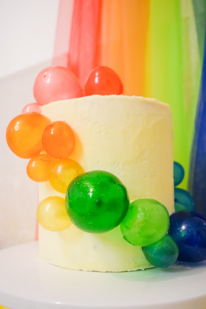  a white coloured cake with rainbow coloured spheres cascading down the sides and a rainbow background