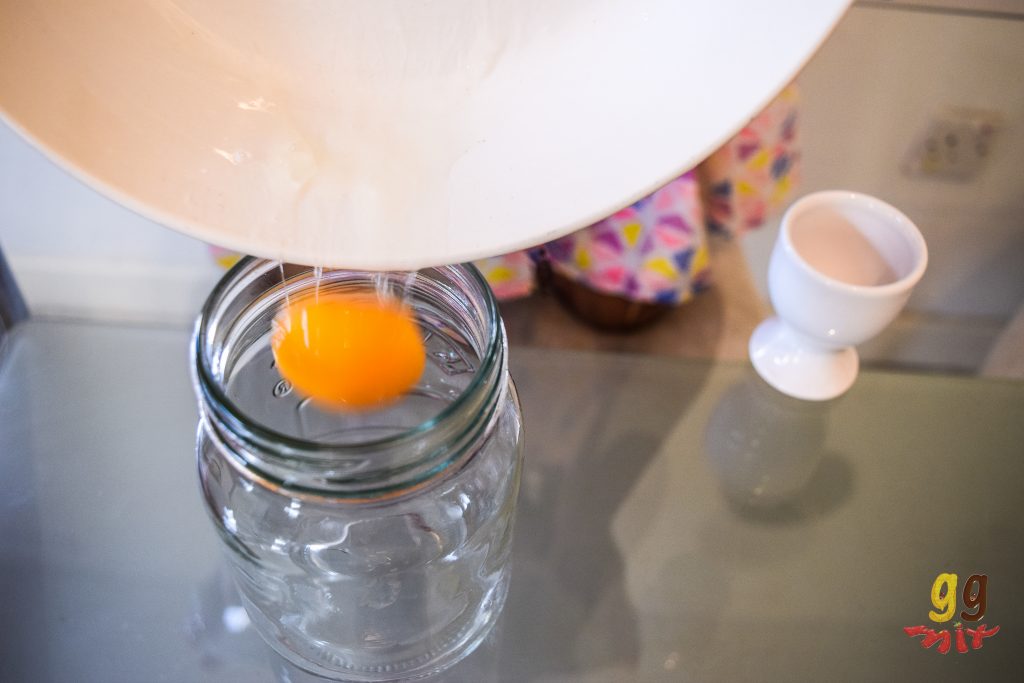 an orange egg yolk is falling out of a white bowl into a glass jar. A white egg cup is to the right of the jar