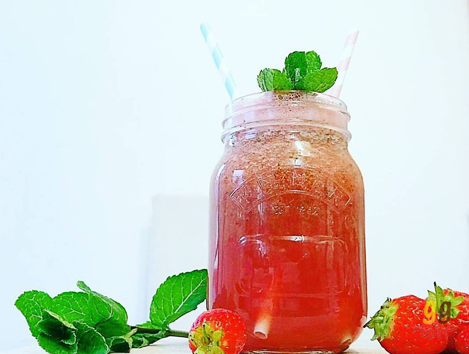 Healthy watermelon, strawberry, mint and rose smoothie drink in a glass jar with sprigs of mint on top and also by its side. Strawberries are also on the other side of the smoothie and a blue and white striped straw and a pink and white straw are in the smoothie ggmix