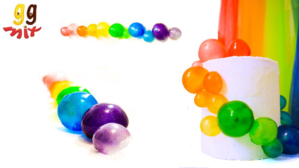 at the top a row of rainbow gelatin bubbles coloured red, orange, yellow, green, blue and purple. Below a a row of rainbow gelatin bubbles balls coloured red, orange, yellow, green, blue and purple coming towards you and a white coloured cake with rainbow coloured spheres cascading down the sides