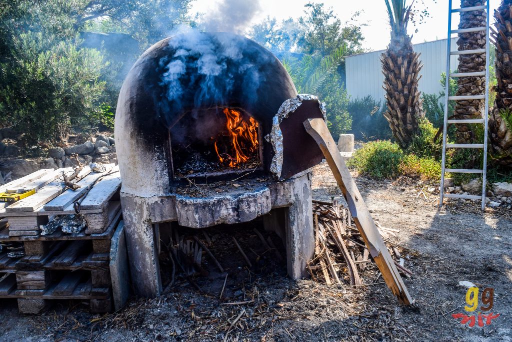 A TRADITIONAL CLAY KLEFTIKO OVEN WITH THE DOOR OPEN WITH FLAMES BURNING INSIDE AND SMOKE COMING OUT OF IT