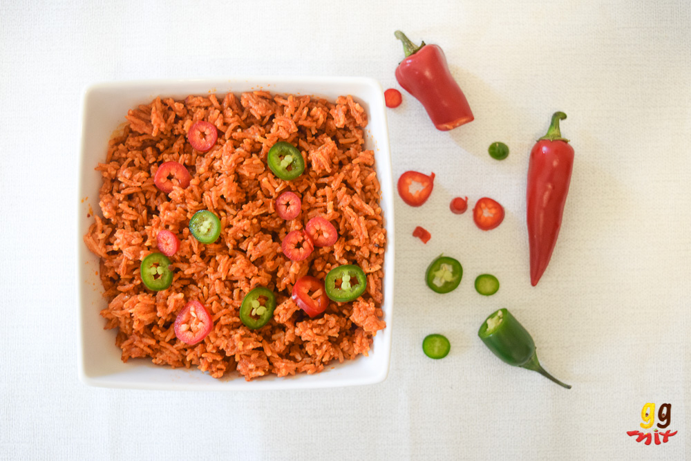 ghanaian jollof rice in a bowl with sliced red and green chillies sprinkled on the top on the side are 1 1/2 red chillies, 1/2 a green chili and slices of red and green chilllies