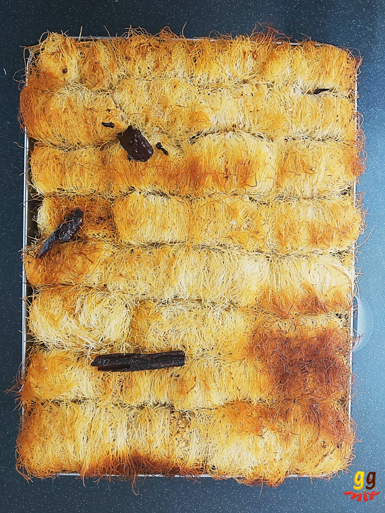 a tray of 32 golden cooked kataifi rolls with some cinnamon sticks and cloves on top